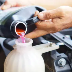 Eine Hand gießt rosa Kühlflüssigkeit aus einem schwarzen Behälter in einen weißen Ausgleichsbehälter eines Autos.