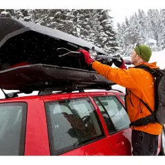 Mann lädt Skier in eine offene Dachbox auf einem roten Auto im Schnee.