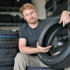 Mann mit roten Haaren hält einen Autoreifen, im Hintergrund Reifenstapel.