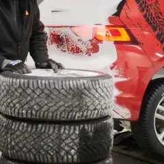 Person mit Handschuhen stapelt Winterreifen neben einem roten Auto mit Schneespritzern und Reifenprofil-Aufklebern.