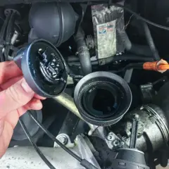 Hand hält abgenommenen Ölfilterdeckel über offenem Ölfiltergehäuse im Motorraum eines VW T5.