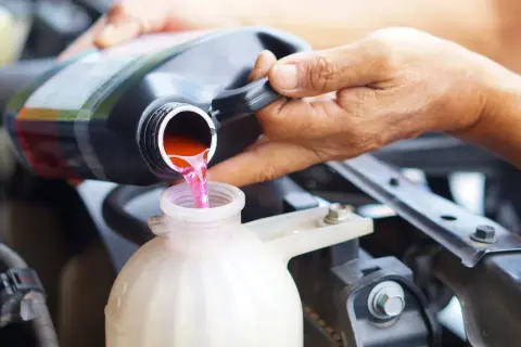 Eine Hand gießt rosa Kühlflüssigkeit aus einem schwarzen Behälter in einen weißen Ausgleichsbehälter eines Autos.