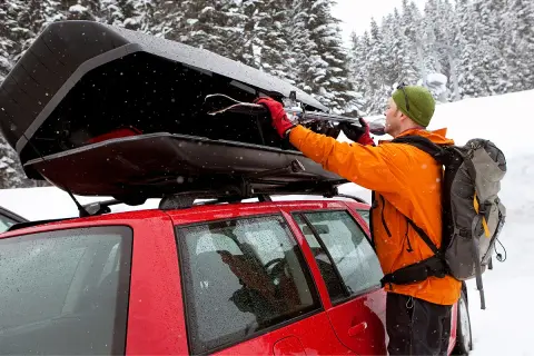 Mann lädt Skier in eine offene Dachbox auf einem roten Auto im Schnee.