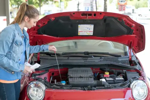 Junge Frau prüft mit einem Ölpeilstab den Motor eines roten Autos.