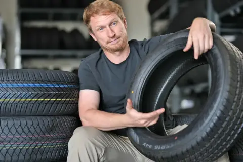 Mann mit roten Haaren hält einen Autoreifen, im Hintergrund Reifenstapel.
