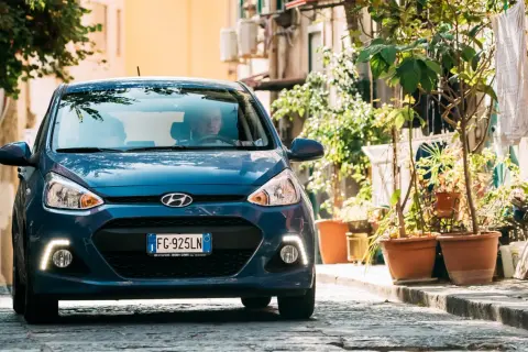 Blauer Hyundai i10 fährt auf einer Kopfsteinpflasterstraße, umgeben von Pflanzen und Gebäuden.