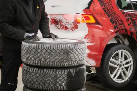 Person mit Handschuhen stapelt Winterreifen neben einem roten Auto mit Schneespritzern und Reifenprofil-Aufklebern.