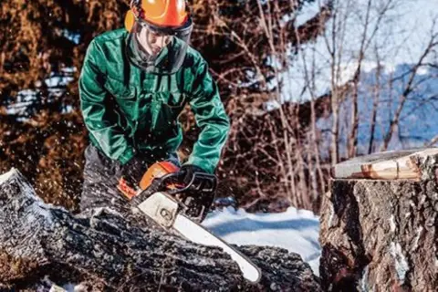 Mann in Schutzkleidung sägt mit Kettensäge in winterlicher Waldlandschaft.