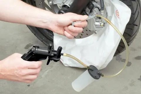 Hand wechselt Bremsflüssigkeit an Motorrad mit Werkzeug und Auffangbehälter.