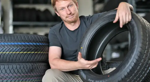 Mann mit roten Haaren hält einen Autoreifen, im Hintergrund Reifenstapel.