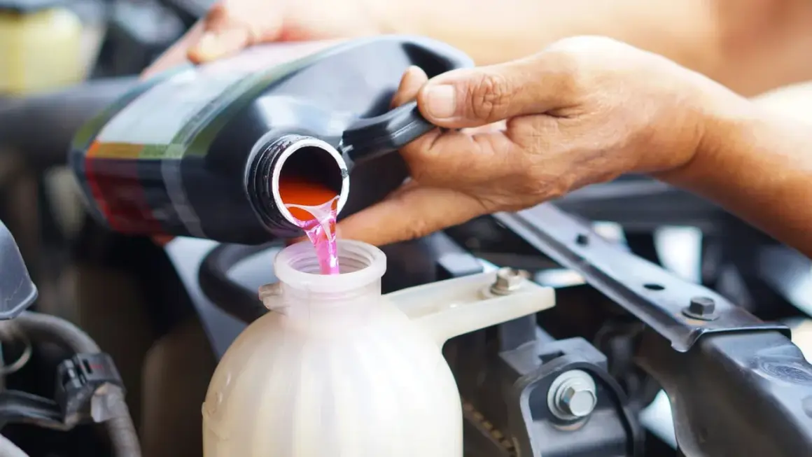 Eine Hand gießt rosa Kühlflüssigkeit aus einem schwarzen Behälter in einen weißen Ausgleichsbehälter eines Autos.