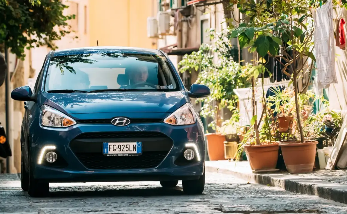Blauer Hyundai i10 fährt auf einer Kopfsteinpflasterstraße, umgeben von Pflanzen und Gebäuden.