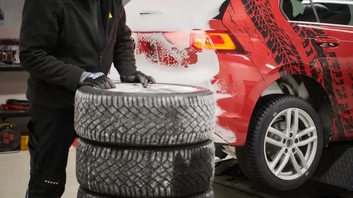 Person mit Handschuhen stapelt Winterreifen neben einem roten Auto mit Schneespritzern und Reifenprofil-Aufklebern.