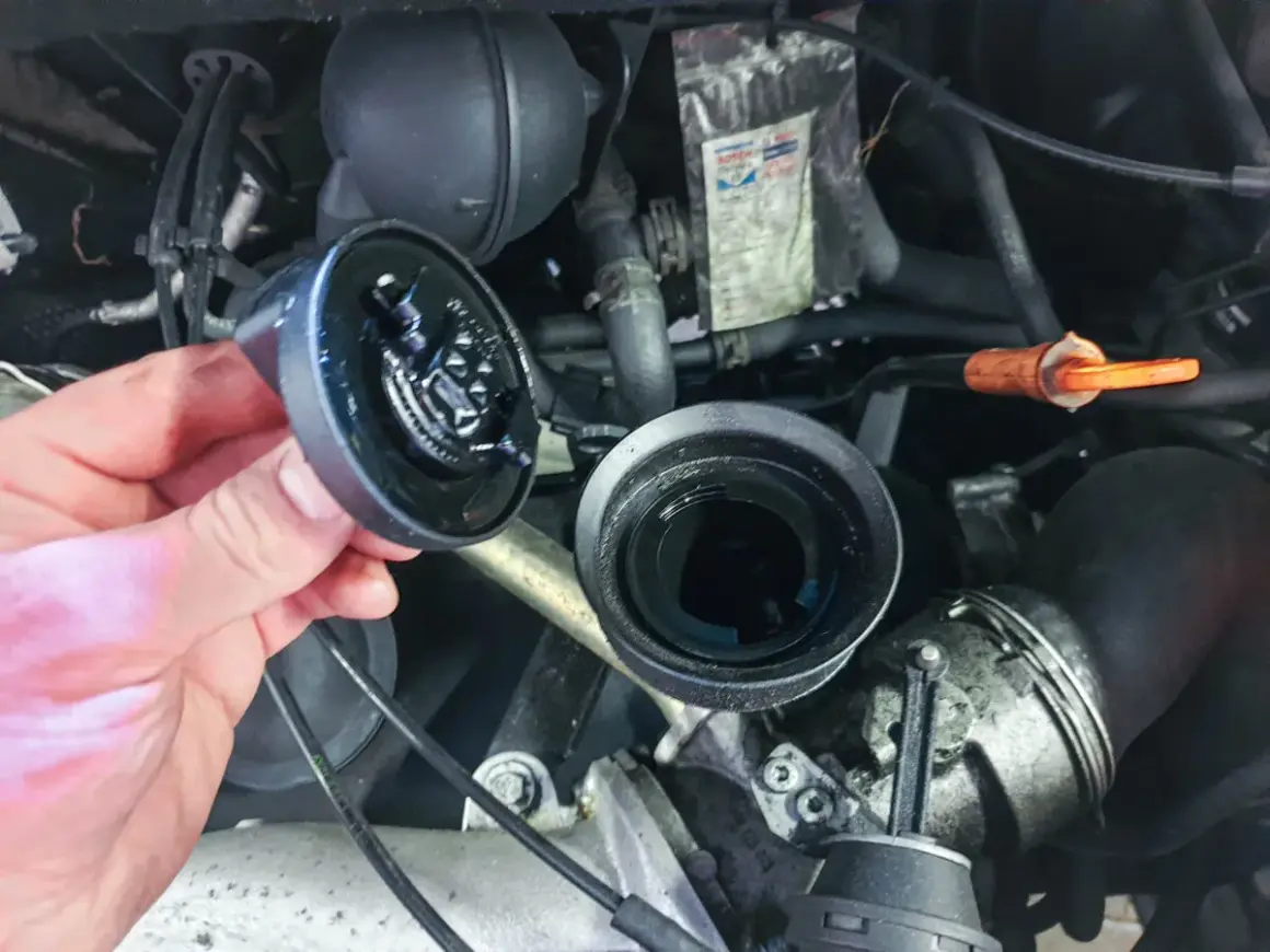 Hand hält abgenommenen Ölfilterdeckel über offenem Ölfiltergehäuse im Motorraum eines VW T5.