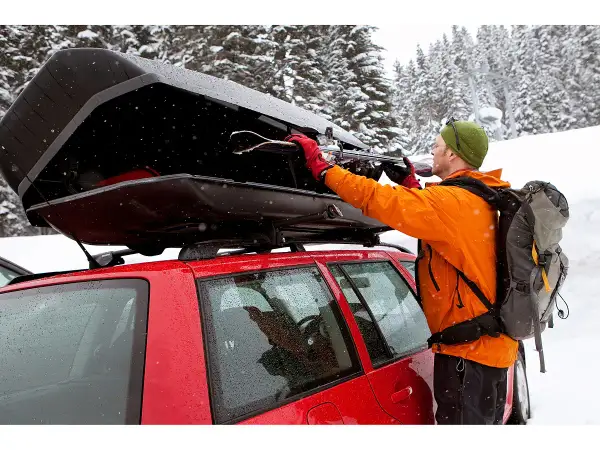 Mann lädt Skier in eine offene Dachbox auf einem roten Auto im Schnee.