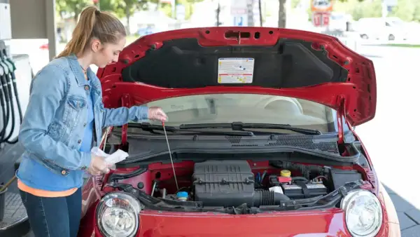 Junge Frau prüft mit einem Ölpeilstab den Motor eines roten Autos.